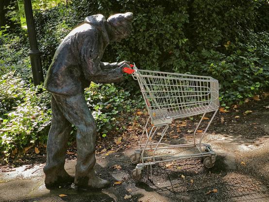 Im Park. Eine Skulptur. Lebensgroße Figur schiebt gebeugt einen Einkaufswagen.