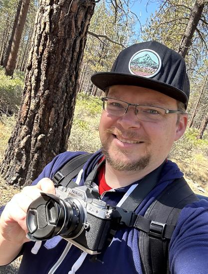 a smiley bearded fellow on the trail, wearing a cap which says Love from the Hood, Portland, Oregon. Camera in hand, with the lens cap on to protect it from all the dust.