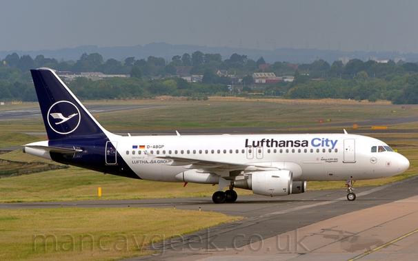 Side view of a twin engined jet airliner taxiing from left to right.
The plane is largely white, with dark blue "Lufthansa City" titles on the upper forward fuselage, the latter word in a lighter blue.
The black registration "D-ABGP" is on the upper rear fuselage, next to small EU and German flags, with additional "Member of Lufthansa Group" text on the lower rear.
The tail and rear fuselage are a dark blue, with the white outline of a circle containing a stylised white flying bird in the middle.
A smaller, blue on white, version of this logo is under the cockpit windows on the plane's nose.
Green grass and black and grey taxiways fill the foreground, as well as a large chunk of the background, leading up to trees and houses in the distance, under grey-blue skies.