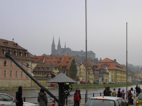 Cars, tourists and a river in the foreground. In the background are city buildings and, further back and above, the spires of the Dom towering over the town. The sky is overcast and the Dom is slightly obscured by fog.