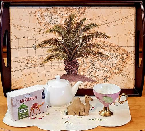Tea tableau in sandy tones with delicate hints of blush pink and olive green. The backdrop is artwork of a palm tree superimposed over an ancient map, inset in a rosewood tray. Upon an ecru runner with eyelet cutouts are a plump white teapot, an elegant tall antique gold leaf pedestal cup, a porcelain camel, and a box of Moroccan Mint green tea decorated with a camel in a desert.