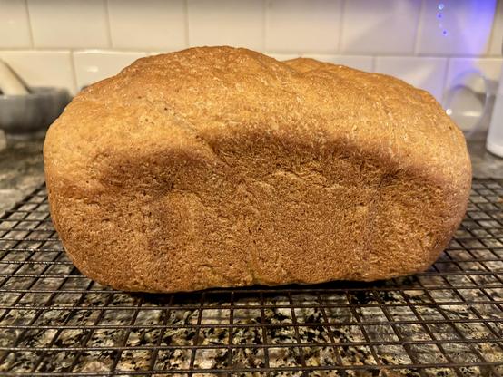 A golden-brown loaf of homemade oatmeal bread cooling on a wire rack. The bread has a domed top with a rich, evenly-browned crust that shows visible oat flecks throughout. The loaf has good height and structure, sitting on a black wire cooling rack against a kitchen counter background with white subway tile backsplash.