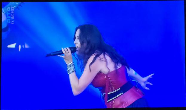 Sharon don Adel profited worth her looking to the left. She's holding her mic in her left hand and had leather looking bracelets on that wrist. She's white. She had long, black hair. She's wearing a drop red bustier. The background is blue