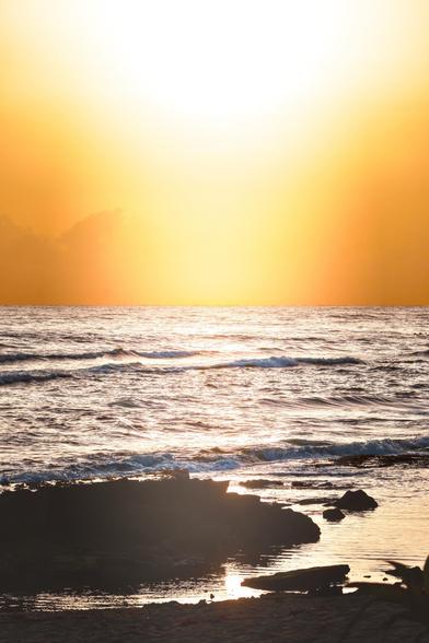 Un sereno paisaje marino al atardecer, con el sol proyectando un cálido resplandor anaranjado sobre el océano. Las olas rompen suavemente contra la costa y se pueden ver formaciones rocosas en primer plano. El ambiente es apacible y tranquilo.
