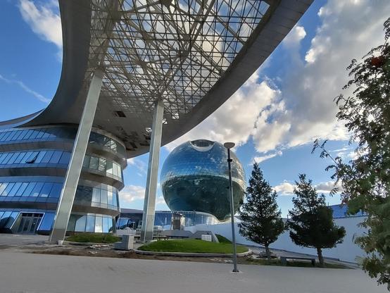 A futuristic mirror-glazed building with a shade canopy above a walkway. In the distance a large mirror glazed spherical building. To right of frame three trees. The conifers and a broadleaf. The sky is blue with white fluffy clouds