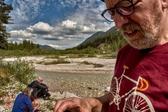 Farbfotografie im Forrmat 3:2 Landscape.
Ein Mann mit kurzem Vollbart, schwarzer Brille und rotem T-Shirt, auf dem ein Rennrad gedruckt ist, steht vor einer Gebirgslanschaft, durch welche ein kleiner Fluss in einem sehr breiten Kiesbett, welches größtenteils trocken liegt, fliest.