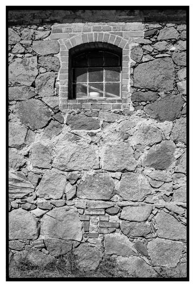 Black and white photograph in portrait orientation. A small window in a rough stone wall with lots of texture.