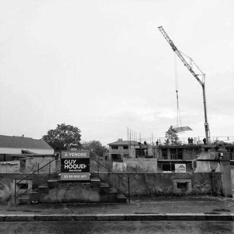 Schwarzweißbild einer Abrissbaustelle am dörflichen Straßenrand. Dominant steht im Vordergrund links der Bildmitte noch eine zweiläufige Hauseingangstreppe, während dahinter schon neue Fundamente gelegt werden. Ein Baukran ist rechts im düsteren Bild zu sehen.