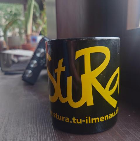 Eine schwarze Tasse mit gelbem Aufdruck des StuRa der TU Ilmenau darauf. Sie steht auf einem dunkelbraunen Fensterbrett aus Holz. Im Hintergrund sind Pflanzen, ein Ordner und ein Radio zu erahnen.