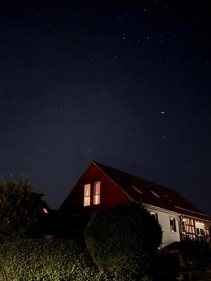 Nachtszene mit einem Haus mit beleuchteten Fenstern, umgeben von dunklem Laub. Der Himmel darüber ist mit zahlreichen Sternen gefüllt, die eine ruhige Atmosphäre schaffen.