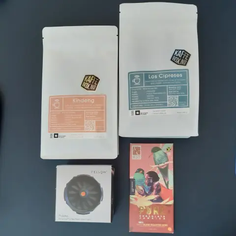 The image features two coffee bags positioned upright, one labeled "Kindeng" in orange and the other "Los Cipreses" in teal. Below them, there is a square box containing the Fellow Prismo attachment for AeroPress coffee maker, which is black with a circular design. Next to the box, there is a chocolate bar wrapped in colorful packaging, labeled "PURE". The items are arranged on a dark surface.