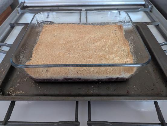 A rectangular glass dish containing a blackberry and apple crumble suits on a baking tray testing on a gas hob. Out of shot the photographer waits impatiently until it is time to taste it.