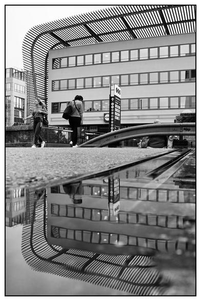 Das Bild zeigt eine städtische Szene in Schwarz-Weiß. Im Vordergrund befindet sich eine große Pfütze am Rande einer Straz, die eine klare Spiegelung der Umgebung erzeugt. Darin sind Teile eines modernen Gebäudes sowie zwei Personen zu erkennen. Eine ist im Begriff, die Straße in Richtung Kamera zu überqueren, die andere geht Richtung Gebäude. Das Gebäude im Hintergrund hat eine auffällige Dachkonstruktion mit parallelen Streben und drei Reihen von Fenstern. An einer Säule ist ein Schild mit Symbolen und Text angebracht, dessen Inhalt jedoch nicht klar lesbar ist.