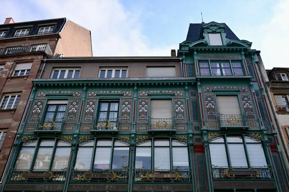 Eine dekorative Gebäudefassade mit architektonischen Details, darunter viele Muster, Blumenmotive, besondere Balkone und großen dreigeteilten Fenstern vor dem Hintergrund eines bewölkten Himmels.