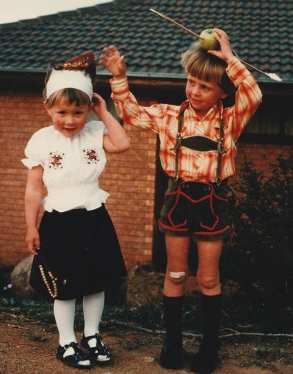 Two young children dressed in European costumes. The boy on the right has an apple on their head pierced by an arrow.