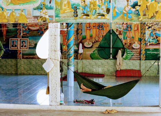 Photo shows a green hammock hanging between some colourful pillars. From the hammock a couple of feet tells of an occupant. A couple of yellow slippers under the hammock of the type used in Buddhist monasteries informs of the occupant’s duties. In the background there is a room with walls on three sides, all painted with religious stories from one-third up the wall. The lowest part of the wall is light green. A large windows opening in the centre of the wall is closed by doors designed as a lotus in deep, green colours. A clothing of sort hangs on the windows handle. Some deep red carpets are rolled together and placed alongside the wall. The floor is lightly cobalt blue.