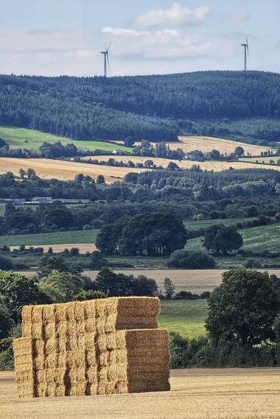 A wide-angle photo of two large, rectangular haybales stacked in a golden field in the foreground, with a rolling Irish landscape of green and brown fields, hills covered in dark forests, and two wind turbines visible in the distance under a partly cloudy sky.