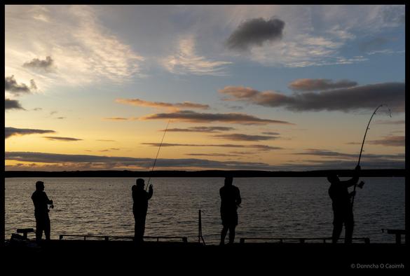 Silhouettes of four anglers fishing from a pier at sunset with dramatic cloudy sky and calm water in Ballycotton, County Cork