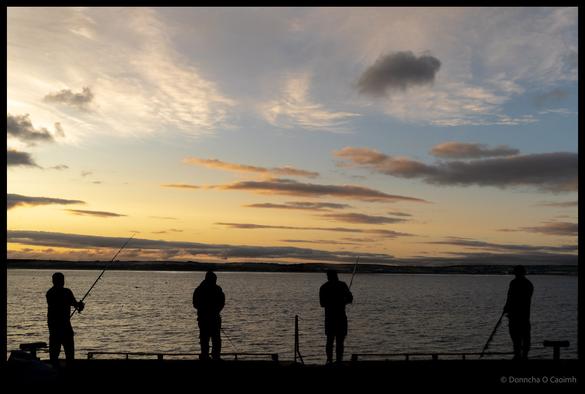Four fishermen silhouetted against golden sunset sky while angling from Ballycotton pier with layered clouds and reflected light on water, County Cork