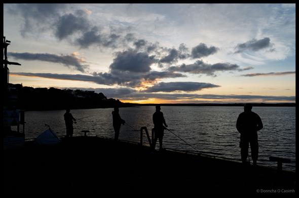 Evening fishing scene showing silhouetted anglers on Ballycotton pier with dramatic storm clouds gathering overhead and village buildings visible in background, County Cork