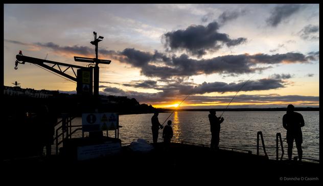 Sunset fishing at Ballycotton pier with harbour crane silhouetted against dramatic sky, anglers casting lines as sun sets over the water, County Cork