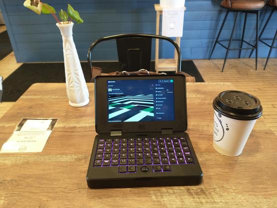 A tiny laptop (MNT Pocket Reform) on a wood grain table at a coffee shop.  There is a paper coffee cup beside it, a white vase on the table behind it, and the blue wooden side of a counter in the background.