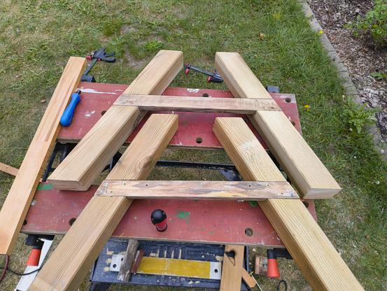 Zwei A-förmige Beine aus Holz liegen auf der Werkbank. In den vorher gesägten Nuten wurde jeweils eine Latte eingelassen. Leider passen die Latten nicht perfekt, denn sie haben oben und unten rund einem halben Millimeter Spiel.
Auf der Werkbank liegen weiterhin ein Handsenker, ein Schraubendreher und unten ein kleiner Hobel sowie eine Alu-Schine, die als Anschlag dient.
