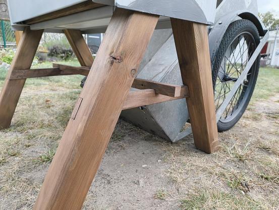 Die fertig montierten Beine an der Bücherkiste. Beide A-Teile stehen parallel zueinander unter der Metallkiste. Zwischen ihren befindet sich eine Quertraverse, die ebenfalls leicht eingelassen ist. Sie wird verhindern, dass die Beine auseinander gehen oder zueinander klappen. Das Holz ist hellbraun lasiert. Hinter der Bücherkiste steht schon ein Handwagen bereit, mit welchen die Kiste morgen früh an ihren Bestimmungsort gebracht wird.