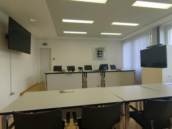 Leerer weißer Gerichtssaal. An der Frontseite das Baden-Württemberg Wappen, links an der Wand ein TV-Monitor, rechts ein Monitor auf einem Podest und von Lamellen verhängte Fenster.

An der Frontseite, unter dem Wappen die weiße Richterbank.
