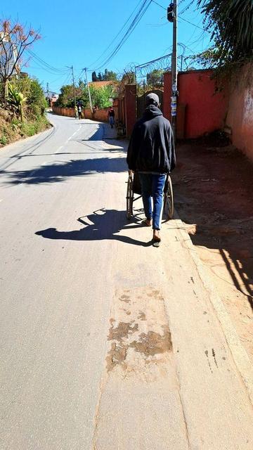 A person walks along the edge of a paved road in Antananarivo, Madagascar, pushing a wheelchair because there is no sidewalk. Cars dominate the street design, forcing pedestrians and people with mobility aids into unsafe conditions.