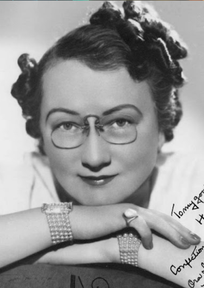 Ora Hanson Snyder, from an autographed headshot; a white woman with dark hair in set curls, wearing pince-nez glasses, matching bracelets and a pinky ring