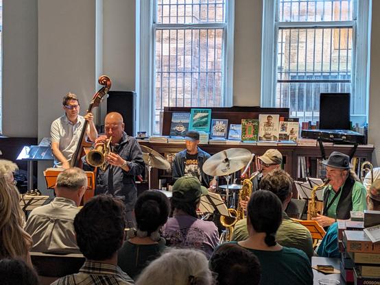 A photo of Steven Bernstein and band performing in Rizzoli Bookstore in New York. Bernstein is playing trumpet at the center of the band, accompanied by upright bass, drums, baritone sax and tenor sax. Several other band members are not pictured. The audience is seated in front of the band. Several books are on display on top of a shelf behind the drummer.