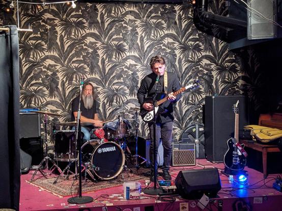 A photo of Galactic Engineers performing live on the small stage of Young Ethel's in Brooklyn. The band is a duo with a guitarist/vocalist and a drummer. Behind the band is a large painting of palm trees in black and white.