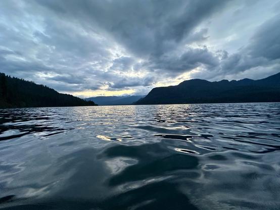 A tranquil lake scene at dusk, capturing calm waters reflecting a cloudy sky with mountains in the background. The light from the setting sun creates a gentle glow on the horizon.