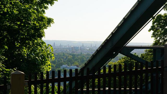 Blick unterhalb der Bergstation, unter der Aufhängung der Bahn durch, ins Tal, Richtung Altstadt 
Gartenzaun im Vordergrund 
In der Ferne sieht man Frauenkirche und co