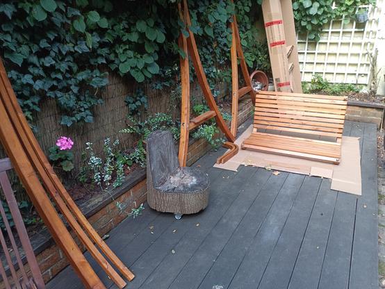 Auf einer Holz-Terrasse liegen Einzelteile eines Holzschaukelsessels, darunter die Sitzfläche und gebogene Seitenteile. Im Hintergrund stehen Kartons, es sind zudem Pflanzen zu sehen und eine mit Efeu bewachsene Wand.