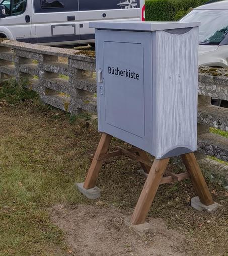 Die Kiste von der anderen Seite aufgenommen. Diese Seite sollte so aussehen, als wären es die Seiten eines Buches. Die Fläche ist weiß übergestrichen und dann streifenförmig abgekratzt. Es ist aber leider nicht so gut zu erkennen.
Im Hintergrund parken Autos.