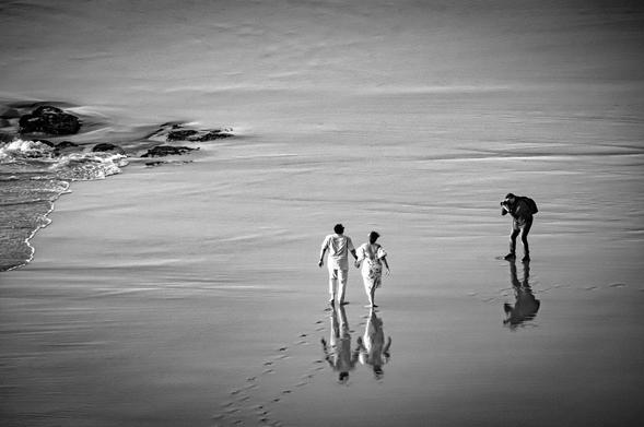 Two people walk on a wet beach, leaving faint footprints in the sand. A photographer captures their moment from a short distance. Waves gently lap against rocks in the background. The scene is in black and white.