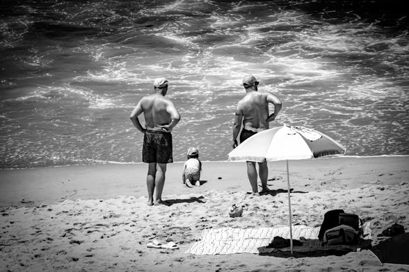 Two adults and a child on a sandy beach, facing the ocean. The adults are standing in swim trunks and caps, while the child crouches near the water's edge. A beach umbrella, cooler, and mat are in the foreground, casting shadows on the sand. The ocean waves appear textured and foamy. The image is in black and white, highlighting contrasts.