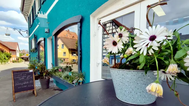 petrolfarbenes Gebäude Krone Randegg, sonniger Blick auf die Längsseite am Schaufenster entlang, überm Eingang ein Kaffeebecher mit Krone, links die Krone am Gebäude eine hängende Krone, viele Blumen vorm Haus