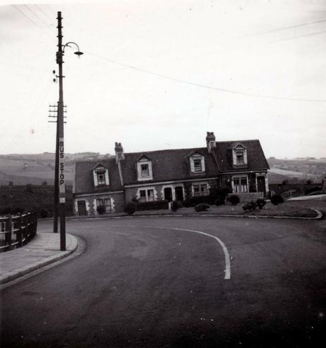 The main feature of this photo is of what looks like the end of a row of cottages that are sloping down to the left. In the foreground is a road that curves to the left and appears to be going down a hill, with a concrete and metal railing fence on the left. In the near foreground is a telegraph pole and lamp post, with the words "Bus Stop" in large letters running down the latter. Behind the cottages is a railway line, and beyond that, a hill.
