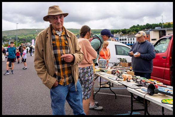A gentleman in tan hat and plaid shirt standing at a busy car boot sale in Bantry with market stalls and shoppers browsing various items spread on tables, County Cork.