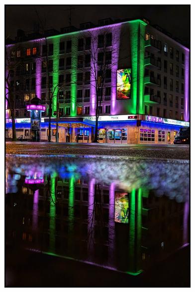 Das Bild zeigt ein mehrstöckiges Gebäude bei Nacht, dessen Fassade in auffälligen Farben beleuchtet ist. Vertikale Lichtstreifen in Grün und Violett verlaufen über die gesamte Höhe des Gebäudes und erzeugen eine lebendige, moderne Atmosphäre. Im Erdgeschoss befindet sich ein Kino, erkennbar an beleuchteten Schildern und Filmplakaten. Die Straße vor dem Gebäude ist nass, vermutlich durch kürzlichen Regen, und eine Pfütze reflektiert die bunten Lichter.