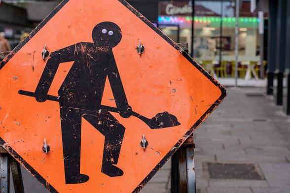 A weathered orange diamond-shaped construction sign showing a black silhouette of a worker with a shovel. Someone has added googly eyes to the worker silhouette's head, giving it a whimsical appearance. The sign has visible wear, scratches, and bullet holes, and is mounted on metal posts. In the blurred background, there are storefronts with colorful neon lighting.