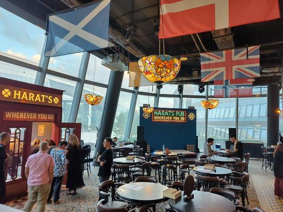 A pub. Large windows in the background, a wooden doorway with the name Harrat's and clover leaf emblems over the door. Flags of various countries hang from the ceiling conspicuously including the butcher's apron or union flag...