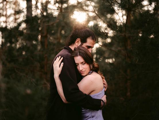 Close-up of a couple standing in a dense pine forest at sunset, their bodies pressed together in a tender embrace during their elopement in Germany, captured by European Elopement and Wedding Photographer Sturmsucht. One partner, wearing a fitted black long-sleeve jacket and sporting dark hair pulled into a neat bun, wraps an arm around the other’s waist and cradles the back of their head. The other wears a strapless lavender gown that drapes smoothly over bare shoulders and down the back, their long, wavy brown hair cascading freely. A simple gold wedding band glints on the right hand resting gently on the partner’s shoulder. Behind them, late-afternoon sunlight filters through tall pine trunks and needles, forming a warm, glowing halo around the pair. Softly blurred evergreen branches in the foreground frame this intimate, secluded moment of quiet celebration.
