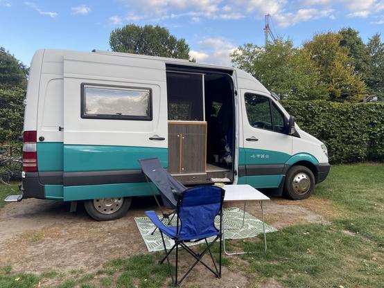 Een witte camperbus met turquoise onderkant in het gras, met de zijdeur open. Ervoor een tafeltje en twee stoeltjes, erachter een heg en bomen