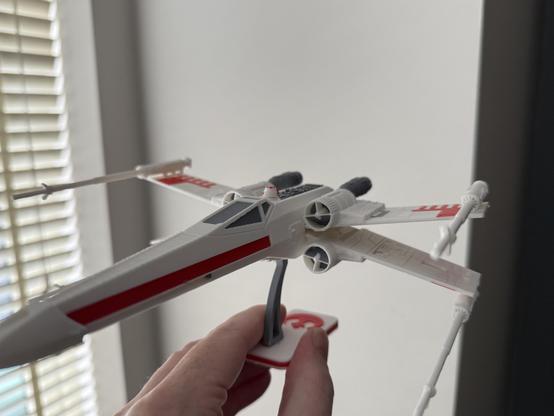 A handheld model of a white X-wing starfighter, featuring red markings and displayed on a stand. Soft light is coming through blinds in the background.