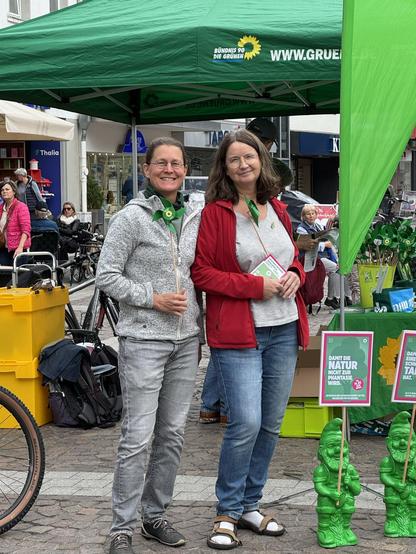 Eine Fotografie von zwei Frauen vor einem Wahlkampfstand der Grünen in Brühl. Beide halten ein grünes Windrad in der Hand. Auf der rechten Seite stehen grüne Gartenzwerge mit Schildern in der Hand. Auf einem Schild kann man den Satz "Damit die Natur nicht zur Phantasie wird" lesen.