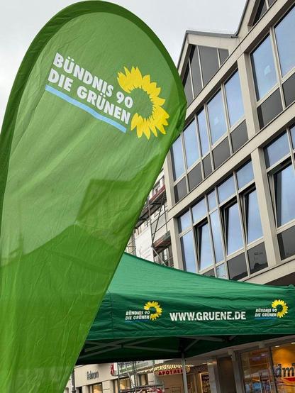 Fotografie einer grünen Beachflag und eines grünen Pavillons der Grünen in Brühl. Beide tragen das Logo von Bündnis 90/ /Die Grünen. Auf dem Pavillon steht außerdem die Webadresse www.gruene.de. Im Hintergrund sieht man eine Gebäudefassade mit vielen Fenstern.